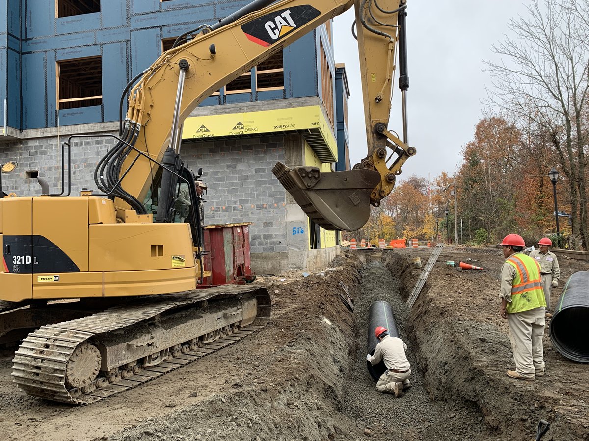 CAT 321D excavator lowering a large concrete pipe segment into an open utility trench while two workers in orange safety vests guide the placement